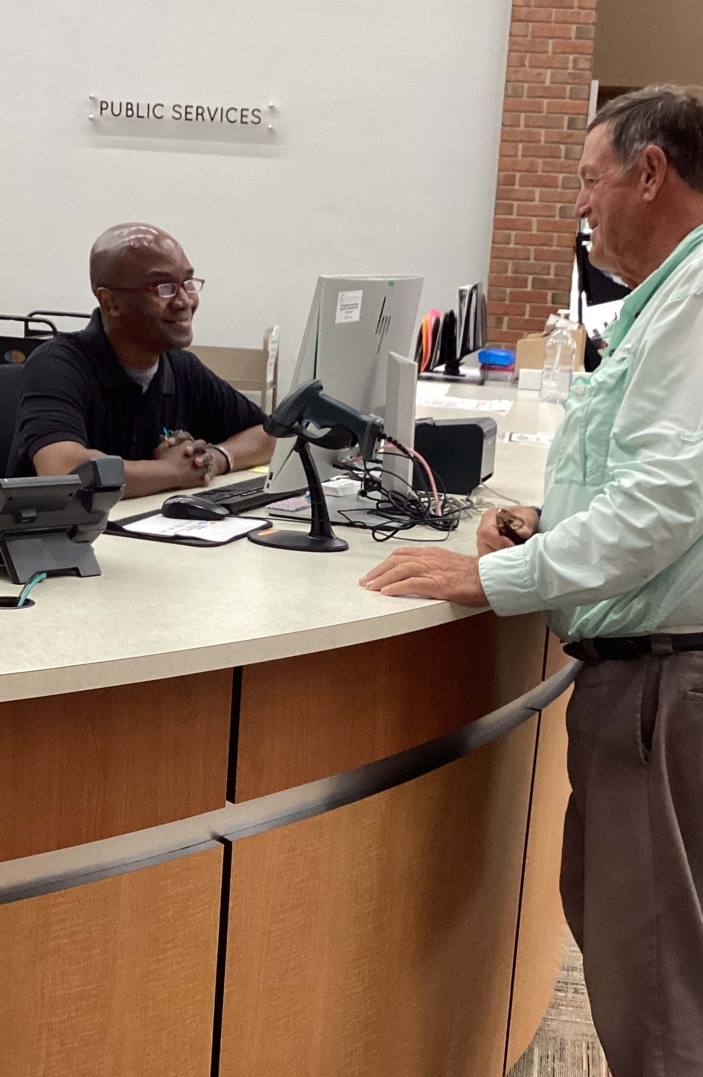 Bald, Black man in glasses sitting behind DPL's Public Services desk, smiling at older white man who standing on the other side of the counter and is smiling back 